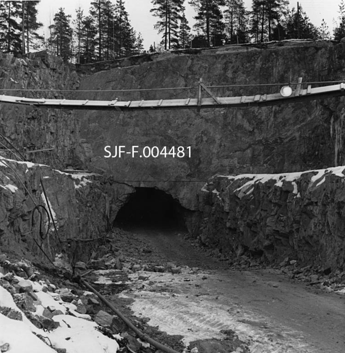 Det søndre innslaget til driftstunnelen for Geithusfoss kraftverk på Modum i Buskerud.  Fotografiet er tatt i januar 1960, fra sørvest, der hvor fordelingsbassenget og kraftstasjonen etter hvert skulle bygges.  Vi ser mot en loddrett nedsprengning i berget med avsatser på sidene, i forlengelsen av tunnelløpet.  Bildet viser også spor etter anleggsmaskiner, som antakelig har hentet utsprengte masser fra tunnelen.  I forgrunnen, over anleggsvegen inn mot tunnelen, hang det ei primitiv gangbru som ble holdt oppe av jernvaiere. 