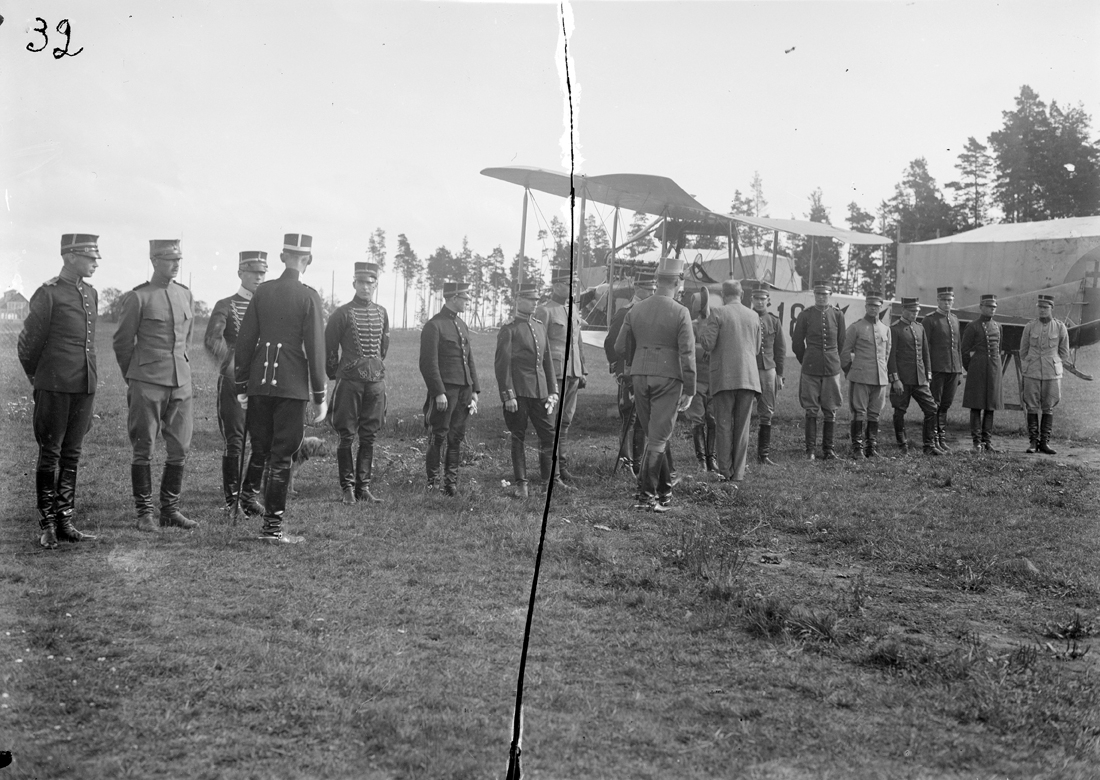 Inspektion av Flygkompaniet på Malmen. 14 st uppställda män och tre män framför. Ett flygplan och en tälthangar i bakgrunden.