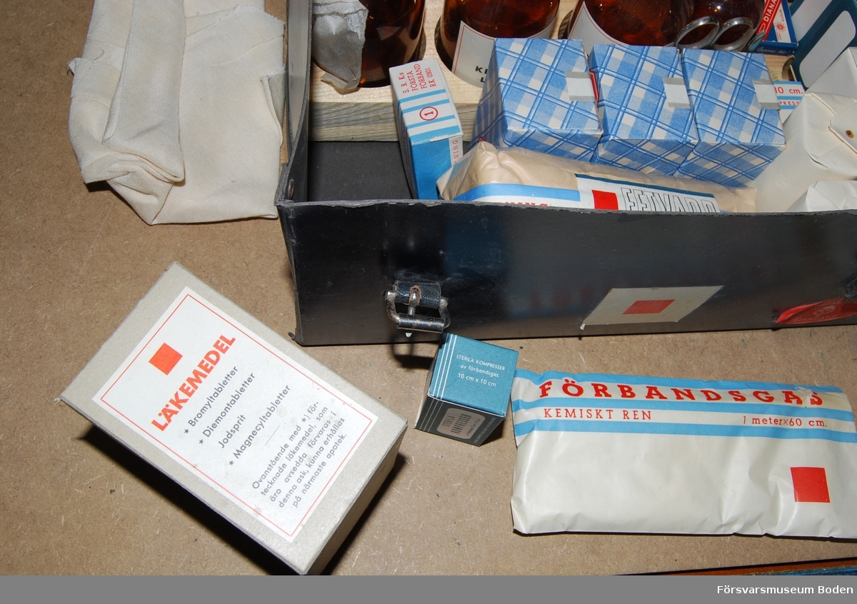 Låda av Unica-papp för förbandsartiklar och läkemedel. Inga läkemedel medföljer. Innehåller också Luftskyddsinspektionens skrift om luftskydd. Innehållslistan på lockets insida är daterad 13/3 1940.