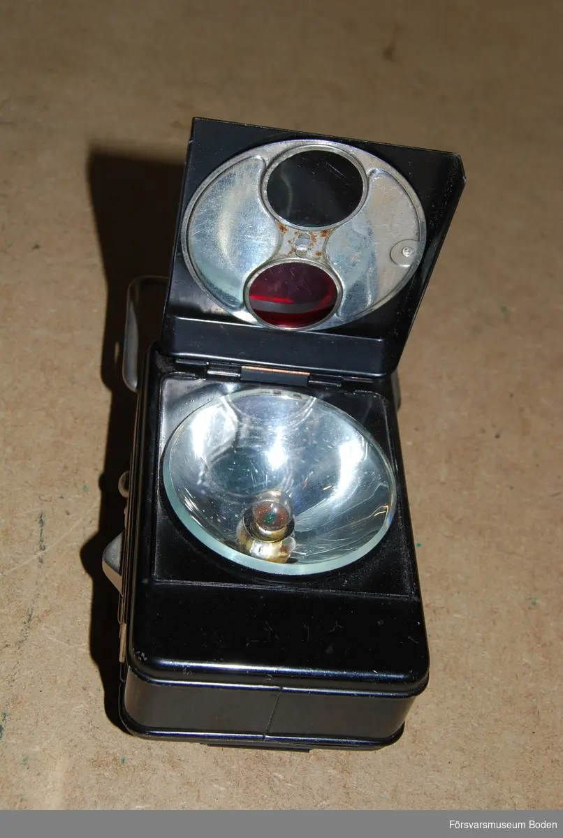 Av svartlackerad plåt, avsedd för två 1,5 V batterier. Uppfällbart lock med grön och röd filterskiva som kan vridas över glödlampan. Av- och påslag genom skjutreglage på sidan. Med tre kronor-stämpel.