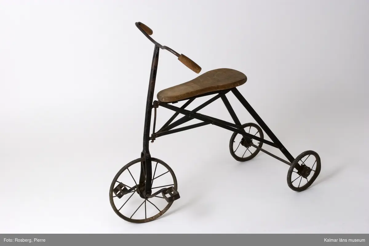 KLM 39704. Cykel, barncykel, trehjuling. Ram av järn, handtag och sits av trä, hjul med massiva gummiringar, trampor på framhjulet.