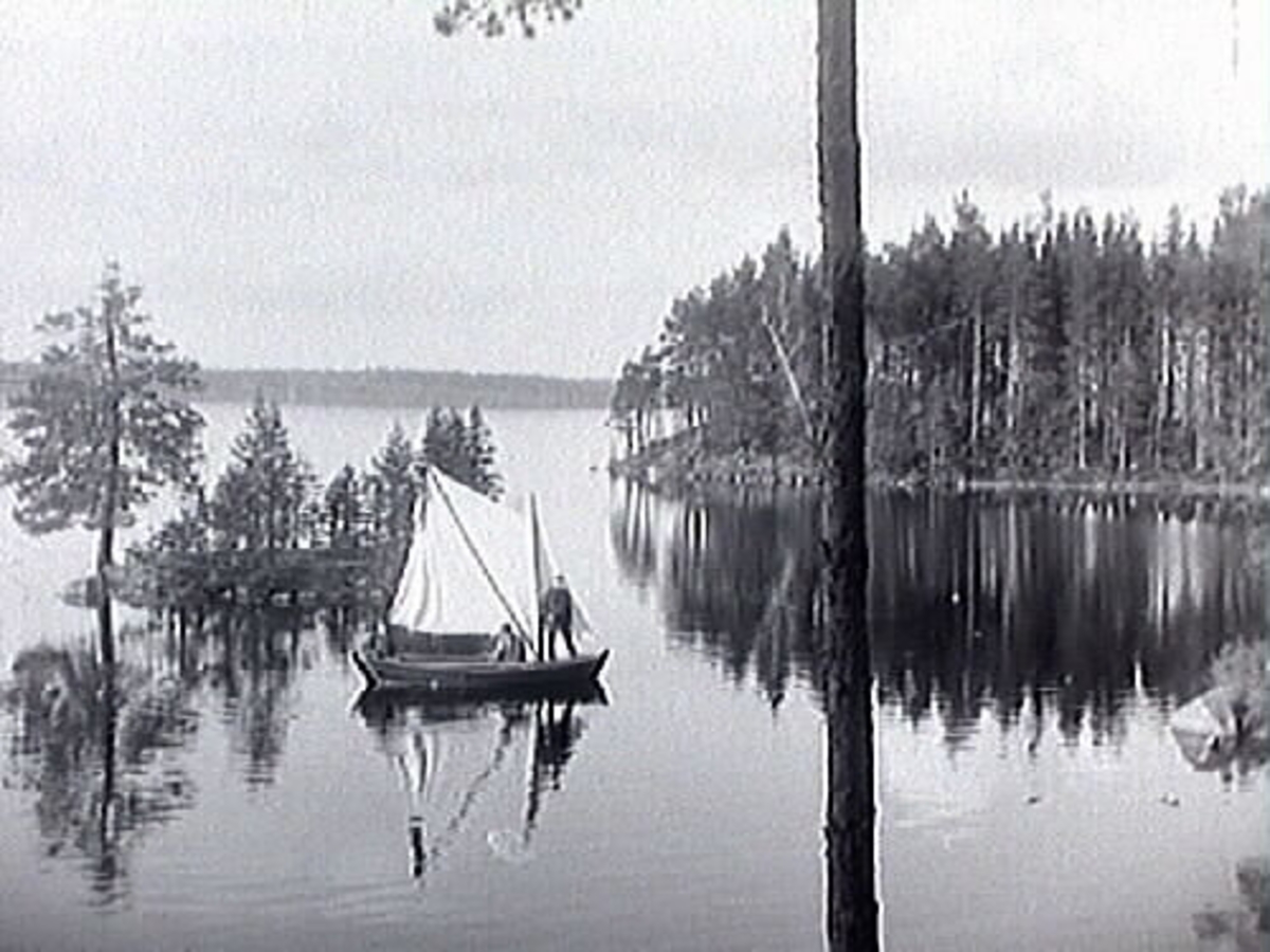 Ett par män i en segelbåt på sjön Hunn i Rejmyre. Sjön är spegelblank. I sjön finns en liten ö med några träd.