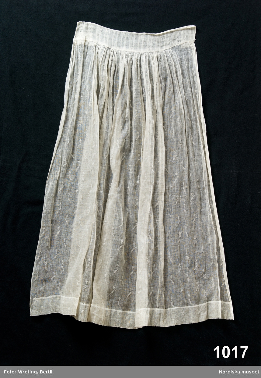 Förkläde av halvt genomskinligt tunt bomullstyg, s.k. linong med ett invävt mönster. Nedtill ca 5 cm bred fåll, upptill rynkat mot 6 cm bred och 40 cm lång linning av linong i annat mönster. Stadkant i sidorna.

Sådana tyger vävdes vid textilfabriker i Skottland i slutet av 1700-talet och kallas "lappet weaving" . Det gjordes med en speciell "lappet frame" en trälist lika bred som varpen och med en rad isatta nålar med öga i änden. Den satt under varpen och styrdes via en excentrisk skiva med mönstret inprogrammerat. Vissa av varptrådarna gick genom dessa ögon och när ramen fördes uppåt så att nålarna stack upp genom varpen och dessutom rördes i sidled genom skivans påverkan så bildade dessa varptrådar ett enkelt mönster på ytan . Mönstret ser ut att vara gjort med ett inplockat inslag, alltså i horisontell riktning men åstadkoms alltså med varptrådar som bringas att röra sig i "fel" riktning. 

Hör ihop med en hel bruddräkt inv.nr. 1012-1018, enligt traditionen använd på midsommardagen 1795 av bruden Annika Andersdotter, i Tunge socken, Ale hd, Västergötland.
Även brudgummens dräkt finns se inv.nr 1019 - 1021.
/Berit Eldvik aug 2005
Om förklädet verkligen bars redan 1795 är det ett mycket tidigt exempel på lappet weaving i bomull. 
/Berit Eldvik 2013-05-15