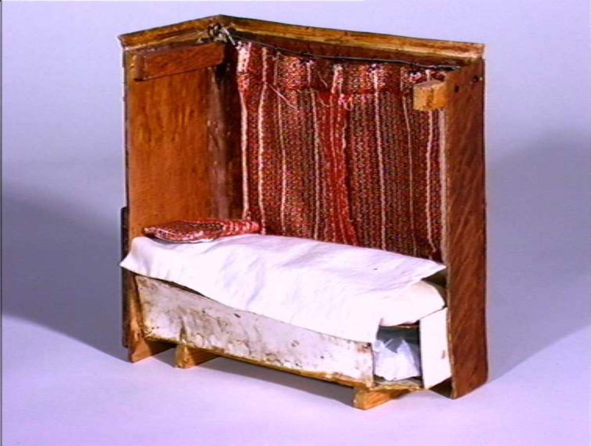 Dukkestue i form av svensk hytte med påmalte tømmerstokker, grunnmur og takstein i tegel. En vegg kan løftes bort og taket kan også åpnes. Stuen har tre stk vinduer med påmalte blysprosser. Inne i stuen er peis, bord og en dukke, en man i svensk folkedrakt med fele, fastmontert. På veggen henger et kurbitsdekorert maleri av Carl Johan.