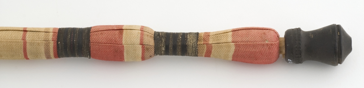 Lang, smal pipe i tre med hode og munnstykke i svart leire, dekormalt med gullfarge. Skaft kledd i tekstil i forskjellige farger.
