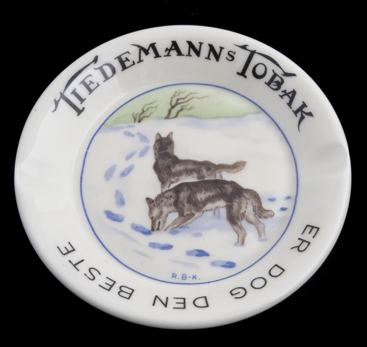Lite, rundt askebeger i porselen med to fordypninger i randen. Påtrykket motiv av to ulver i vinterlandskap etter original av R. B-K.