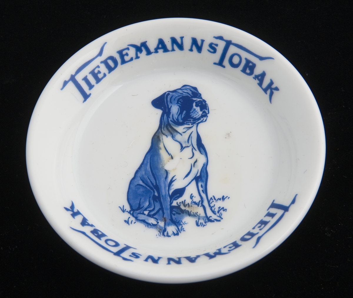 Liten, rund skål i porselen med reklame for Tiedemanns Tobak. Påtrykket motiv i blått av en sittende hund.