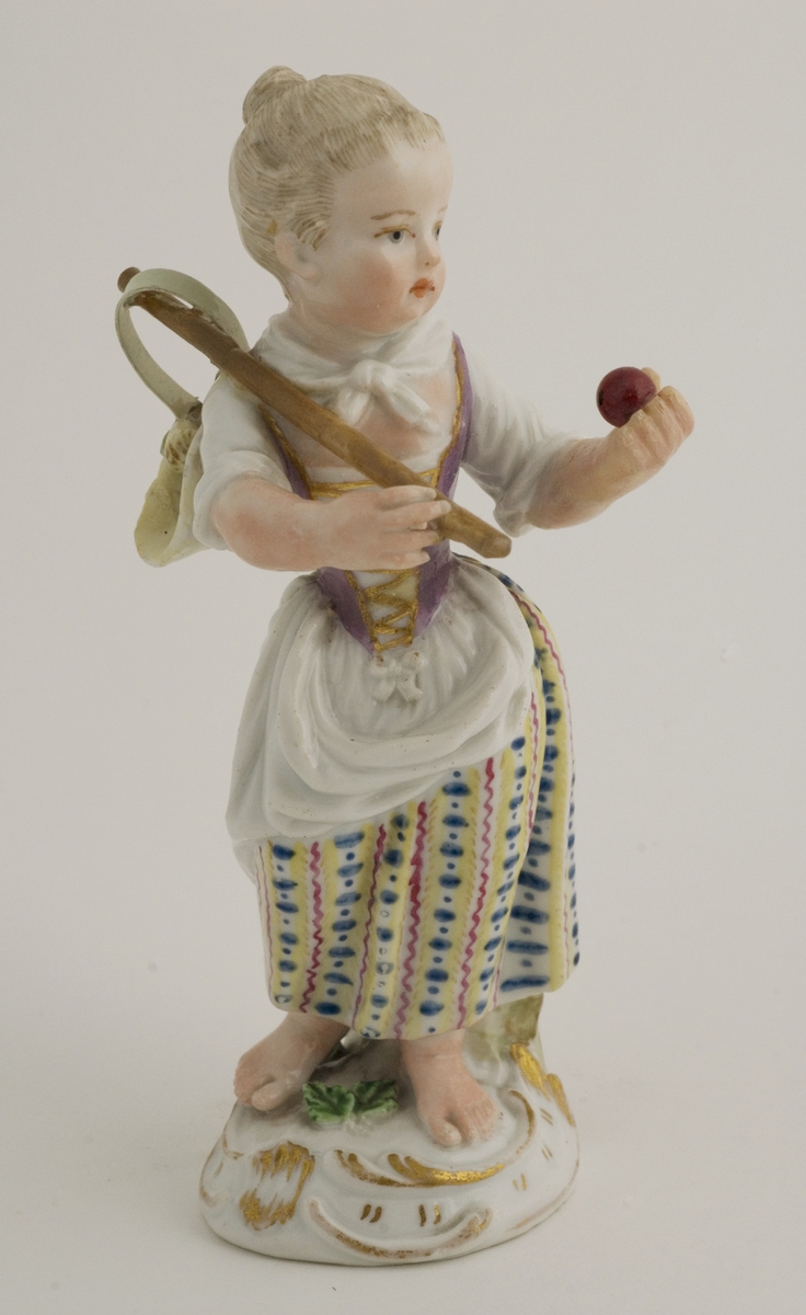 Figur; porselen, pike i stripet skjørt, violett snøreliv, bærer en stråhatt med frukt på stokk over skulderen. Merket 11 med blått under fotplaten.