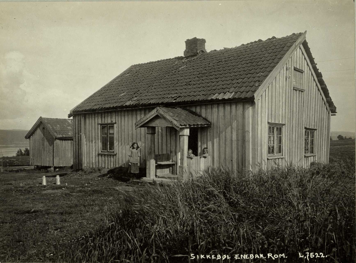 Sikkebøl, Enebakk, Nedre Romerike, Akershus. Lite våningshus med uthus ved siden av. To barn sitter på verandaen, jente står ved siden av. Melkespann på tunet.