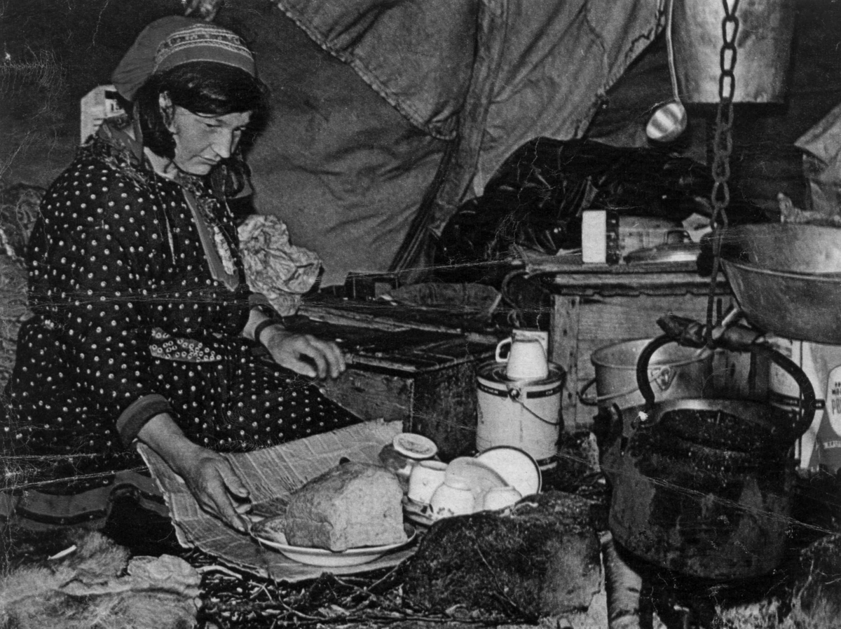 Interiør fra sametelt. En kvinne setter frem mat ved et ildsted med kaffekjele over. Kautokeino. Fra A magasinet, Aftenposten 22. august 1964.