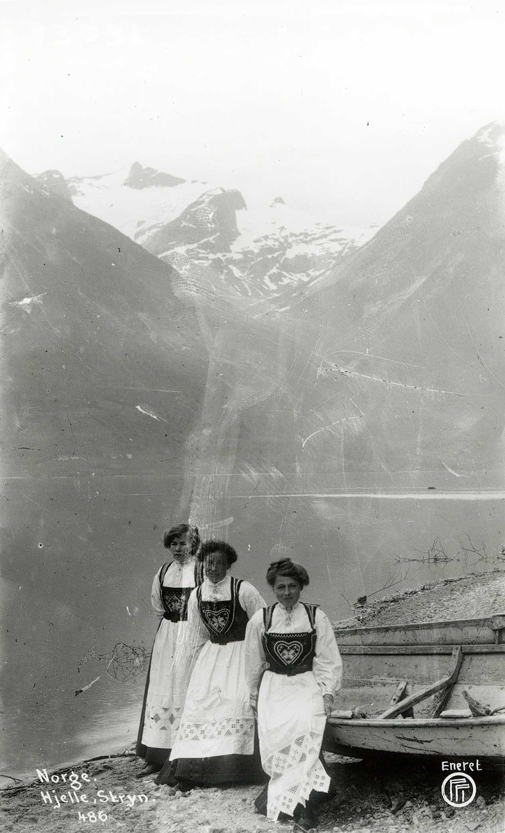 Tre kvinner står foran båt ved fjord med bratte fjell. Drakt fra Hardanger.
