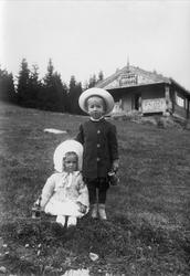 Barn stående i helling, hus i bakgrunnen, ukjent sted.
Seri