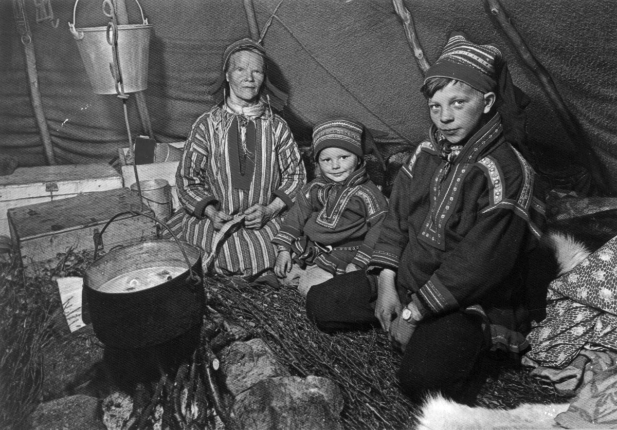 Interiør i et sametelt, tre personer sitter ved kokestedet, en gryte med mat henger over ilden. Finnmark.