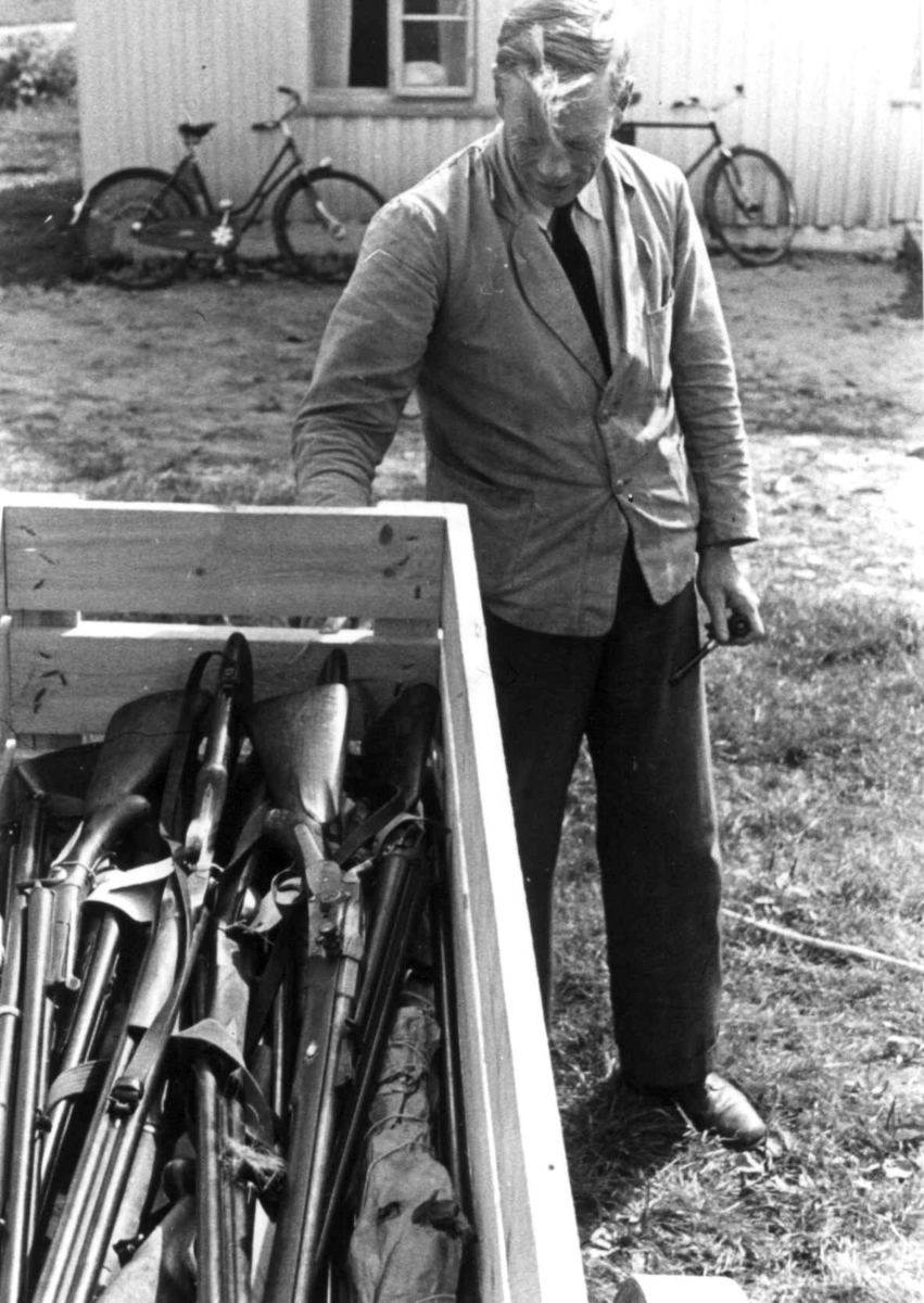 Knut vatnestrøm med en samling våpen i en kasse. To sykler lent mot en husvegg i bakgrunnen. Frostemyr 1941.