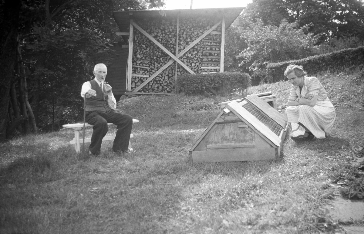 Eyvind Arentz sitter i hagen på Nøtterø 1944. Dordi Arentz betrakter et kaninbut. I bakgrunnen ses et vedlager.