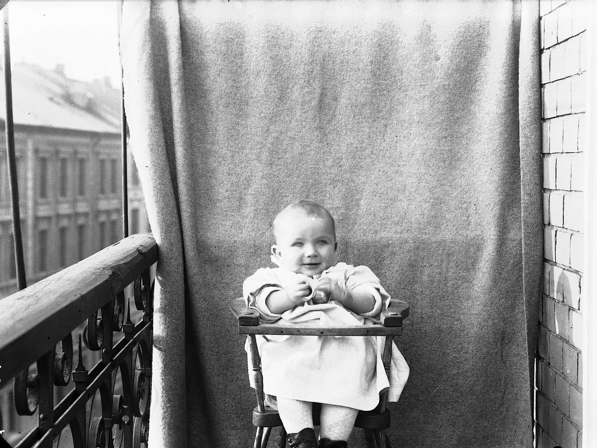 Lillebror Axel Q. Wiborg i barnestol på balkong, ant. Meltzers gate 9, Oslo, 1899. Fotografert mot opphengt bakteppe.
