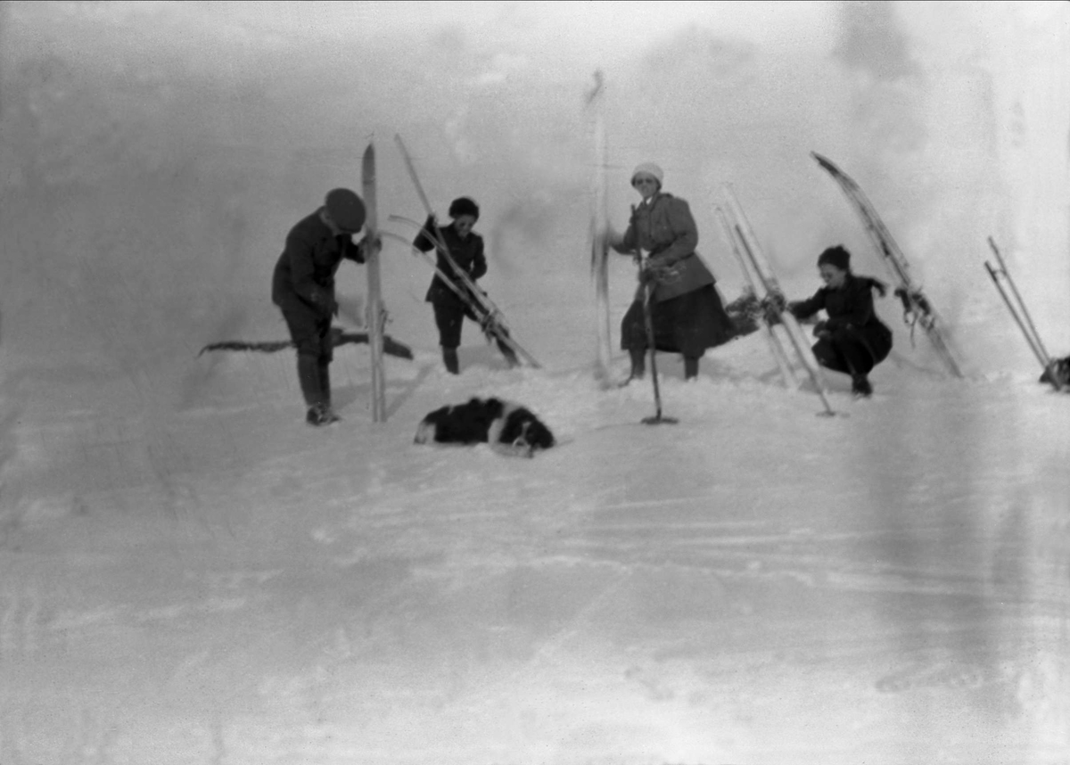 En gruppe mennesker setter fra seg skiene på et snødekket landskap. En hund ligger på bakken. Robsahm og Lund.