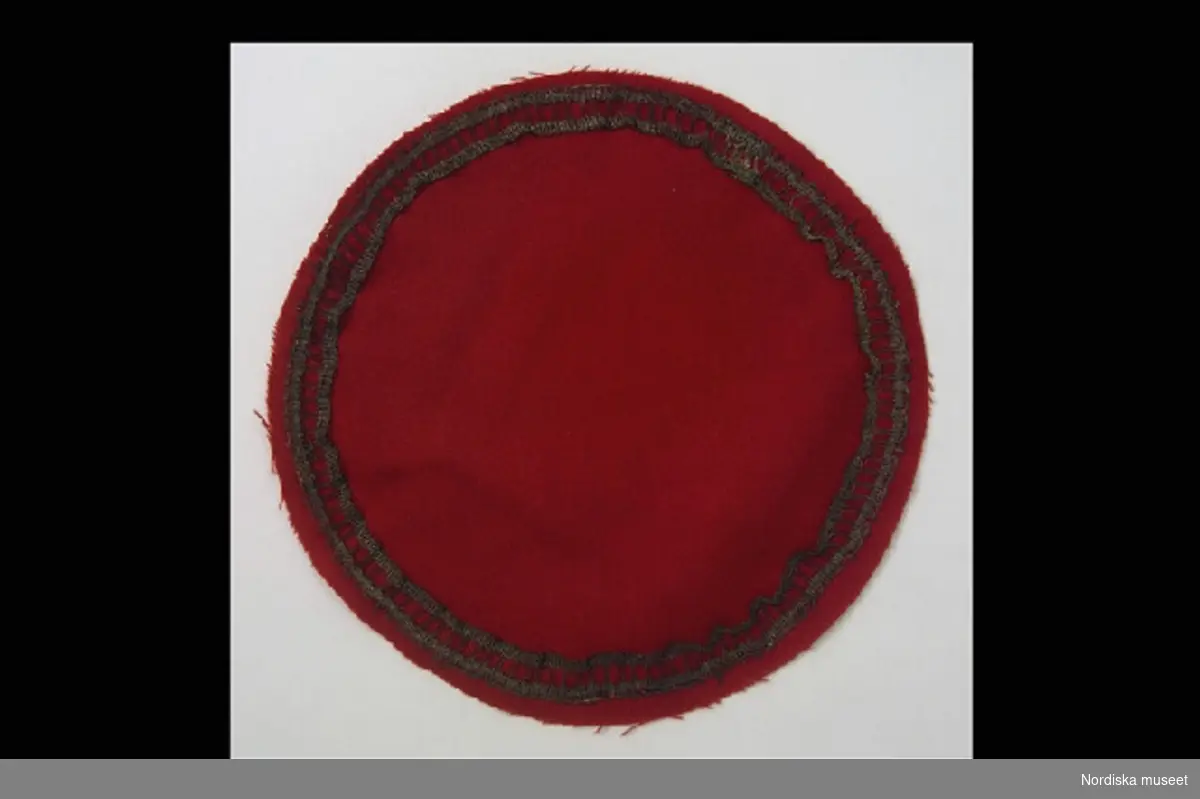 Katalogkort:
"Bordduk, dockbordduk. Rund bordduk av rött kläde/ylle. Dekorerad med metallspets av metallan runt en silkeskärna. HC 1997." [= Helena Carlsson, inventering Sesam 1996-1999]
