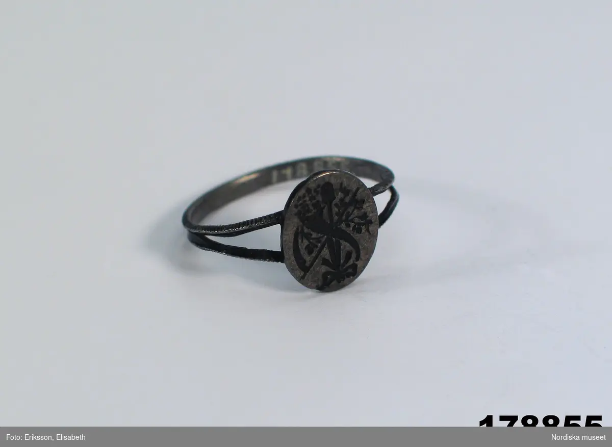 Ur katalogtext från utställningen Smycken, Nordiska museet 1949: 138. Ring av järn, ovalt framstycke med graverade symboler, ett ymnighetshorn, en spade, en lie m.m.