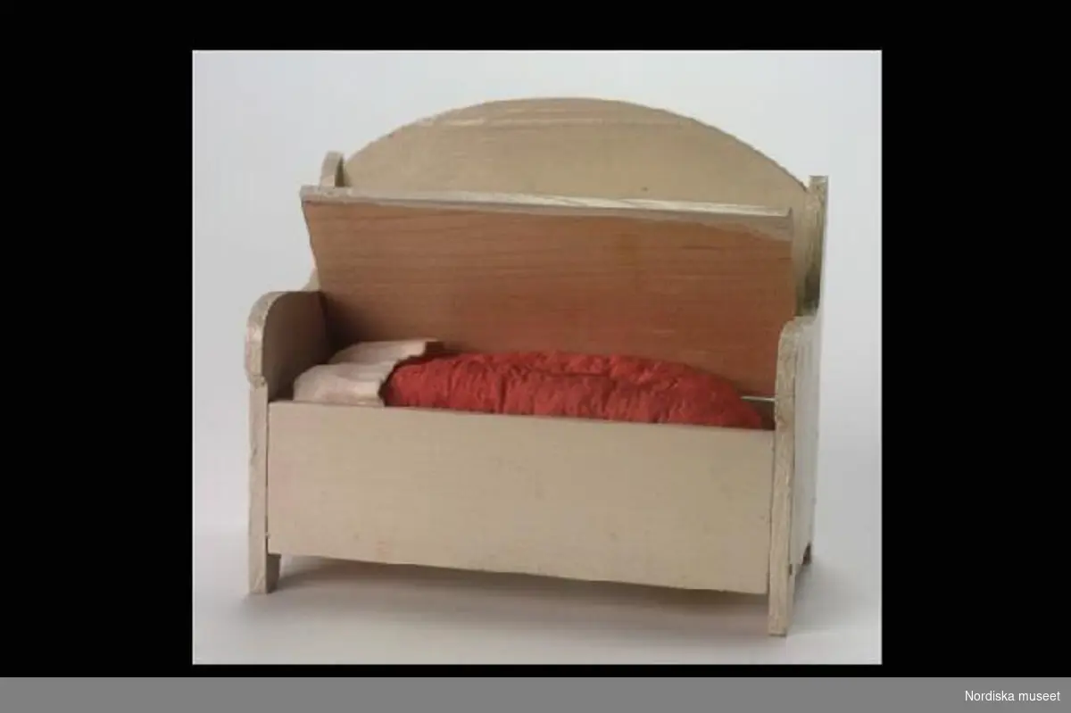 Inventering Sesam 1996-1999:
Bäddsoffa; L 12 cm, B 6 cm, H 11,5 cm
Sängkläder, L 4,5-11,5 cm
Bäddsoffa med sängkläder, a-f
a) bäddsoffa av trä, målad i gulvitt. kontursågade gavlar och halvmåneformad rygg. Sitsen är öppningsbar och däri ligger sängkläderna. Undertill märkt med blyerts "E / 1,25" (trol. prisuppgift).
b) madrass, rosa, rektangulär, stoppad med vadd.
c) lakan av vitt linne, kvadratiskt.
d) kudde av vitt linne, rektangulär, stoppad.
e) lakan av vitt linne, kvadratiskt. 
f) täcke av rosa siden, stickning på översidan, ruggad ull på undersidan.
Brukad av Catarina (född 1919) och Anne-Sophie (född 1921) von Heidenstam, döttrar till givaren.
Tillhör dockskåp 220.405 med inv nr 220.406-220.760
Leif Wallin 1996