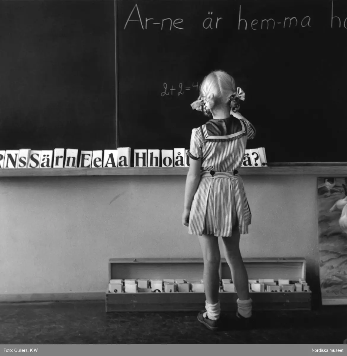 Skola och undervisning. Flicka med margaretaflätor skriver på svarta tavlan i småländsk folkskola.