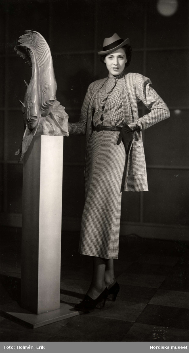 Dammode visas på varuhuset Nordiska Kompaniet i Stockholm 1937. En kvinnlig modell poserar iklädd dräkt brevid en skulptur i föreställande av två fiskar. Kjolen är vadlång, blusen har knytband, jackan är av ledig modell. Till detta bär hon herrhatt, handskar och pumps med klack.