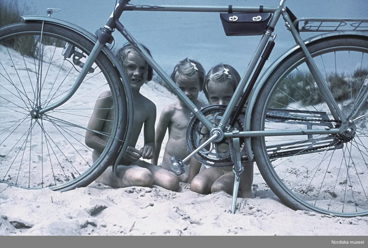 Tre flickor sitter på en sandstrand bakom en cykel.