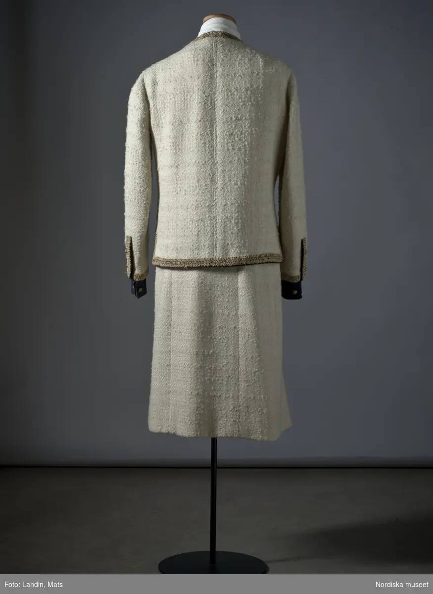Dräkt av vit ylletweed med bandgarnering av svart ripsband och beige-bruna snörmakeriband. Inköpt hos modehuset Chanel i Paris 1961. Nordiska museet inv nr 283341.