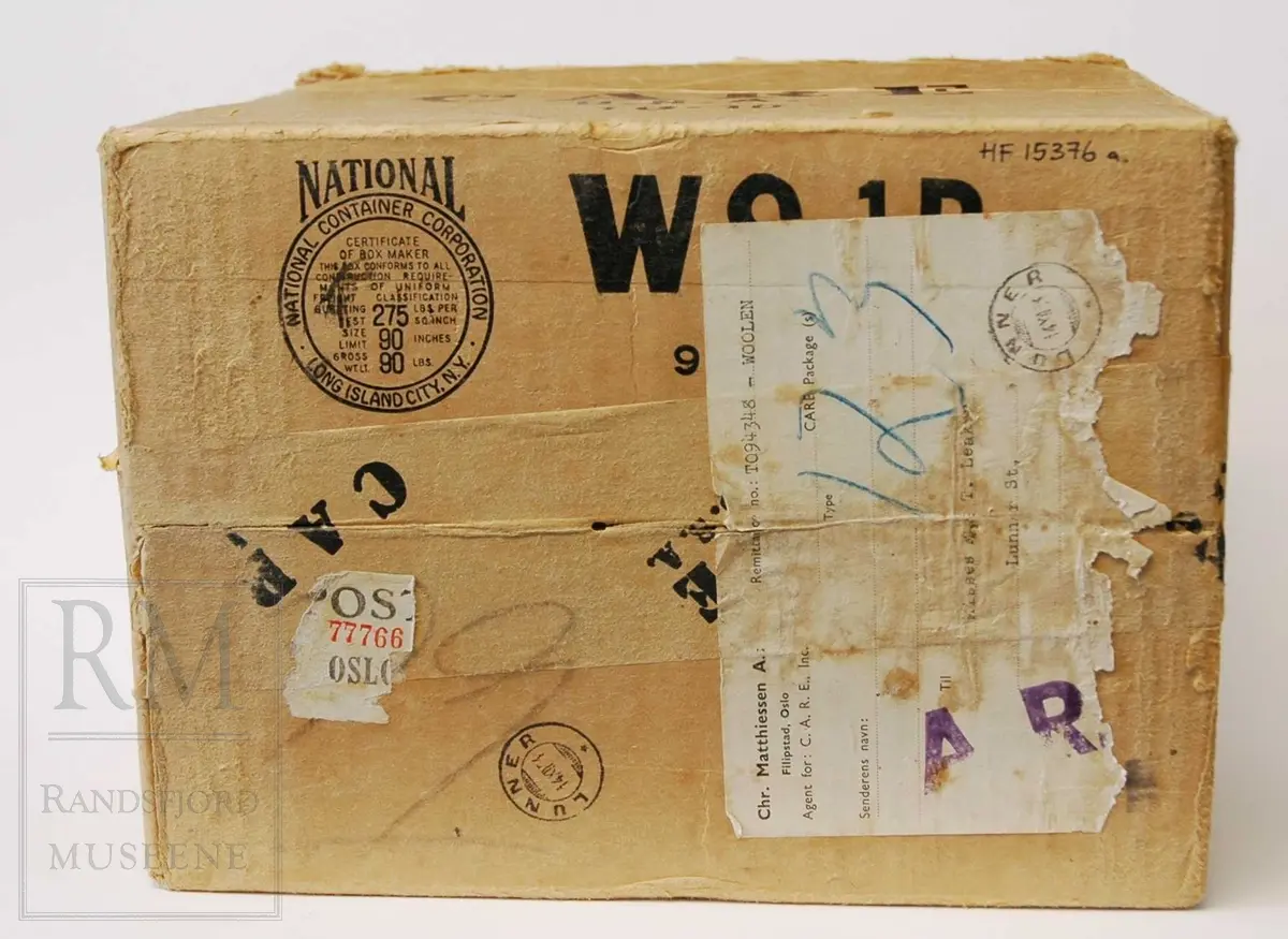 Kvadratisk, avlang form. Pappeske for sending med varer til kriserammede/behovstrengende områder.
Innhold i esken: To pakker med ullstoff. Det ene er i mørk/lys beige: Dreil/ fiskebensvevd kyper. Det andre i ensfarvet brun, samme teknikk.

