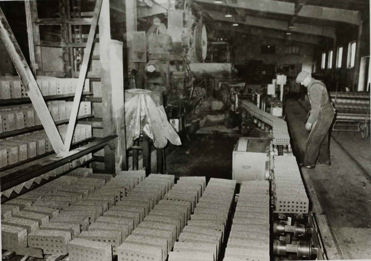 Svend Byberg følger med på et langt samlebånd med murstein i fabrikken til A/S Graverens teglverk. I forgrunnen er det lagret murstein i flere rekker.