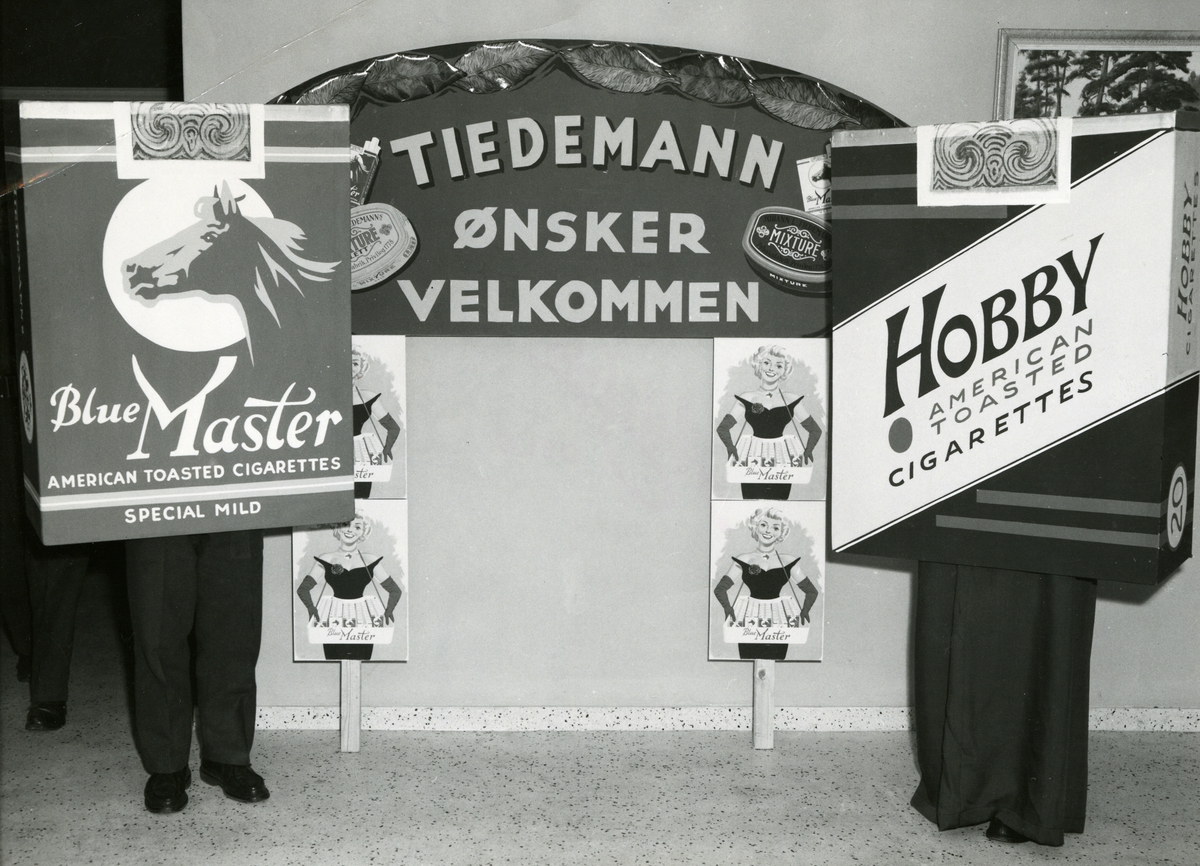 Gratis danseaften på militærforlegning med The Big Chief Jazzband. Tiedemann arrangerte disse aftenene på flere norske militærforleggninger på 1950-tallet. To mennesker utkledde til sigarettpakker, Blue Master og Hobby, foran reklameskilter.