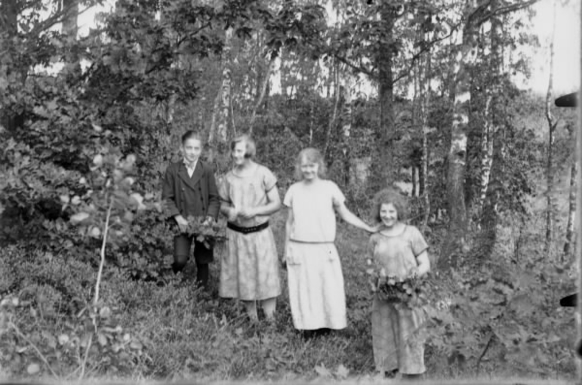 Tre ungdomar, tre flickor och en pojke, står i en skogsbacke med löv- och blomsterbuketter.