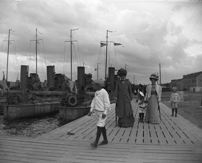Hamn med krigsfartyg, på bryggan går två kvinnor och tre pojkar i sjömanskostymer