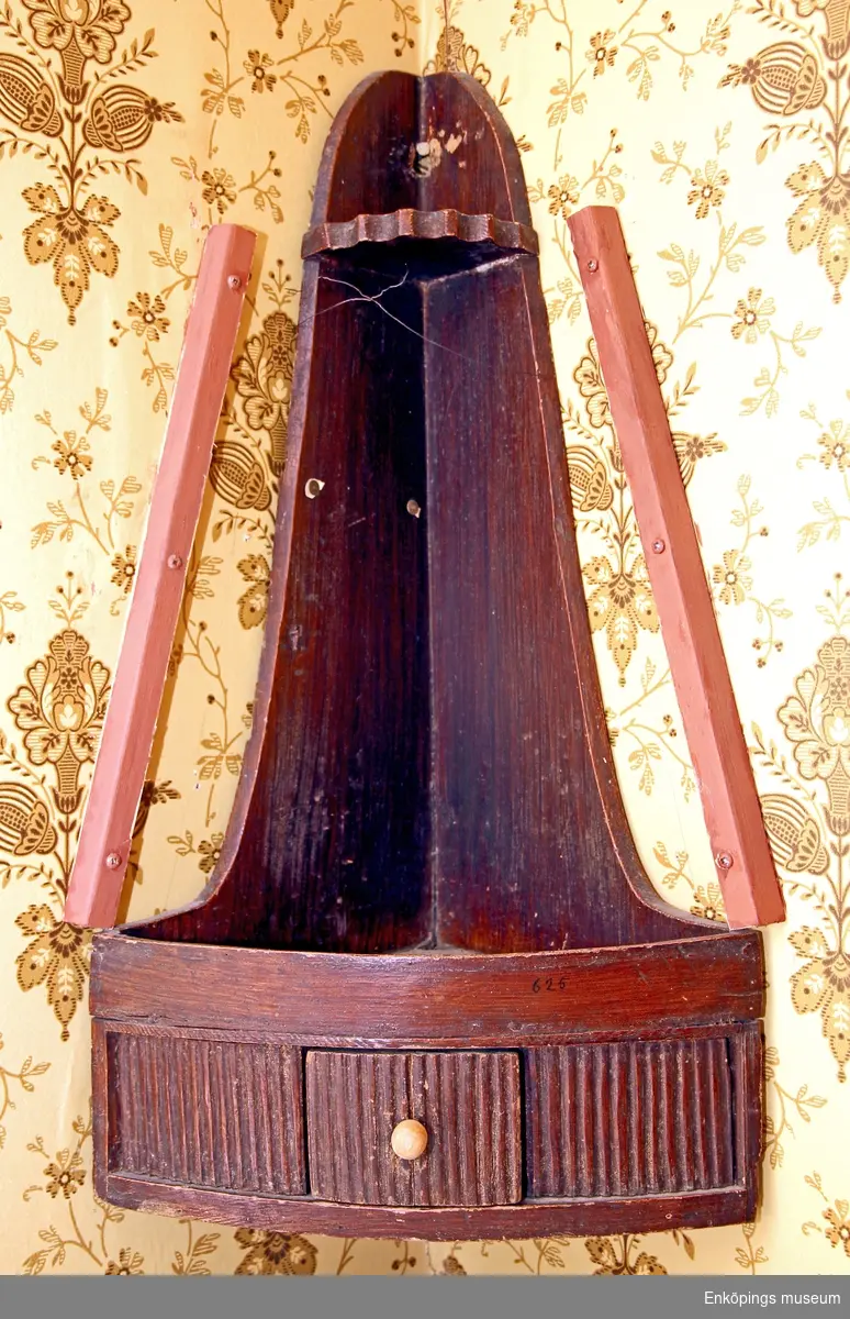 Hörnhylla med triangulär form, avsmalnande upptill, framsidan konvex och delvis räfflad med låda i mitten. Lådan förseddmed knopp av horn, övre delen av pipstället har en hylla med vågig kant, hål för upphängning. tillverkat på 1800-talet.