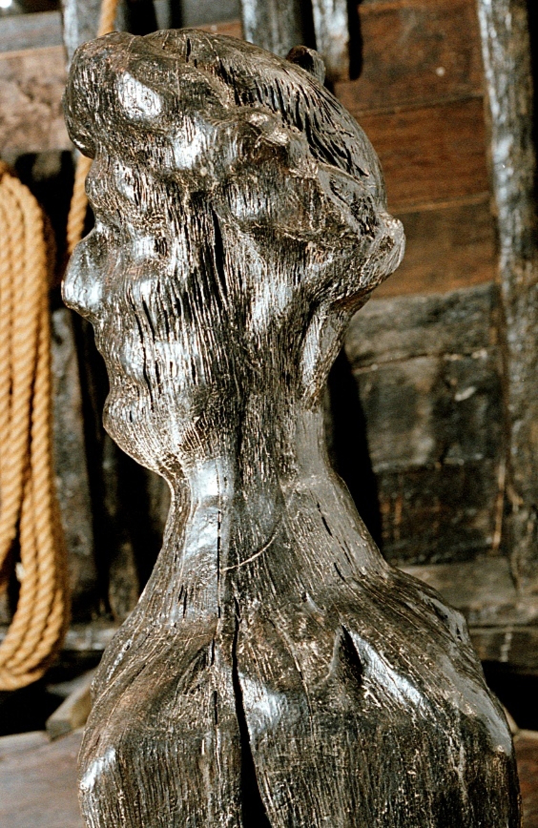 Rektangulär knekt med två skivgatt.
Upptill är knekten krönt med ett tredimensionellt manshuvud med stora mustascher. På hjässan sitter en huvudbonad med bräm och knopp. Runt halsen sitter en dubbelkrage med stora, något spetsiga, flikar. Huvudet är riktat föröver och är snidat i ett med knekten. På knektens sidor finns märken efter stora järnbeslag.
Skulpturen är ytsliten och har några stora, djupa sprickor.

Text in English: A rectangular knight with two sheave holes.
On top of the knight is a three-dimensional male head with large moustaches. On his head is a headgear with trimming and a knob. Round his neck is a double collar with large, somewhat pointed lobes. The head is carved as one with the knight and faces the bow. The sides of the knight have marks from large iron fittings.
The sculpture is surface worn and has a few large, deep cracks.