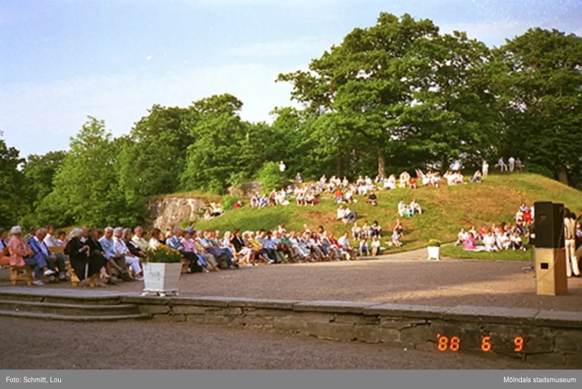 Musikunderhållning av en orkester vid Gunnebo slotts framsida. Människor sitter i gräset och på bänkar.