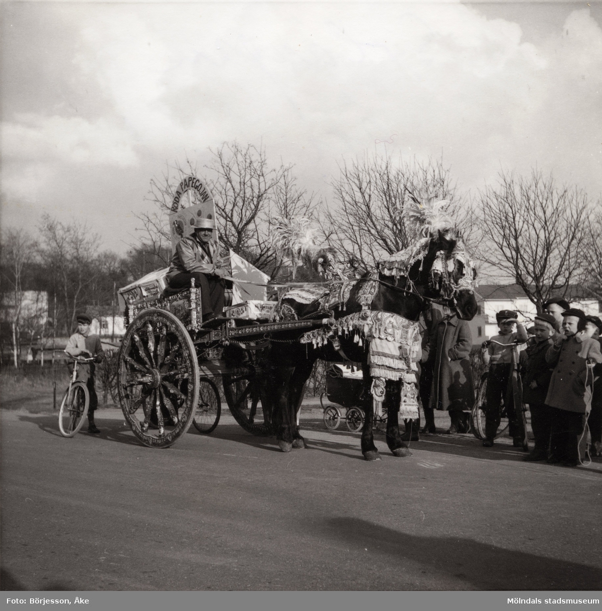 Motiv från Solängen. Häst med vagn och kusk bär dekorationer. Möjligtvis reklam av något slag? Skylt med texten "Röda papegojan" på vagnen. Barn på cyklar står runt omkring och tittar på.
Möjligen Frölundagatan på 1950-60-talet.