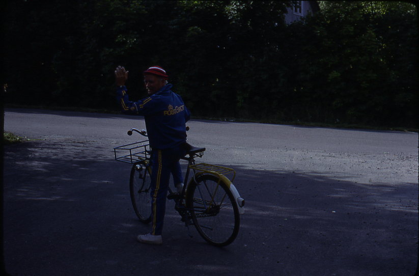 Karl Stenqvist vinkar där han står medd foten på pedalen på sin gula postcykel. Han är klädd i blå overall med gula revärer och Postgirot i gula bokstäver på ryggen. Nära brevbärarexpeditionen Olsborgsvägen i Vallentuna.