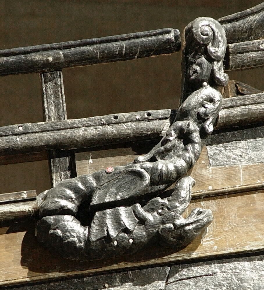 Skulptur av en drake återgiven i höger profil.
Draken har ett stort huvud med kraftigt uppdragen nos och öppen käft. Drakstjärten och vingen övergår i den ovanliggande tritonen, se fyndnummer 23059. Baksidan är slät.
Skulpturen är välbevarad.

Text in English: A sculpture of a dragon in right profile.
Large head with very upturned nose and open jaws. The dragon''s tail and wings change into a Triton that is situated above, see No. 23059. The back is smooth.
The sculpture is well preserved.