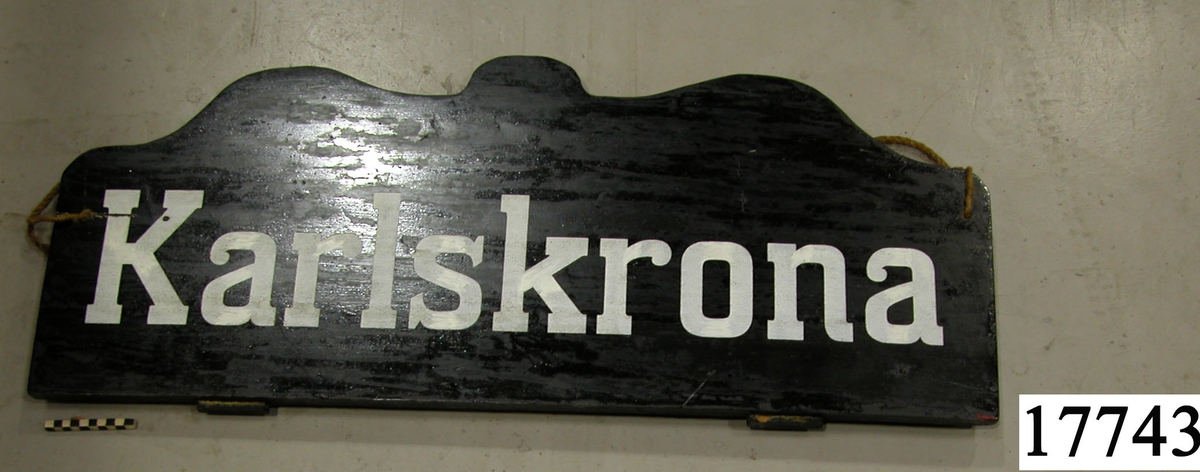 Rektangulär skylt, ovansidan sågad i tre bågar, svartmålad. Vit schablonmålad text: " Karlskrona ".