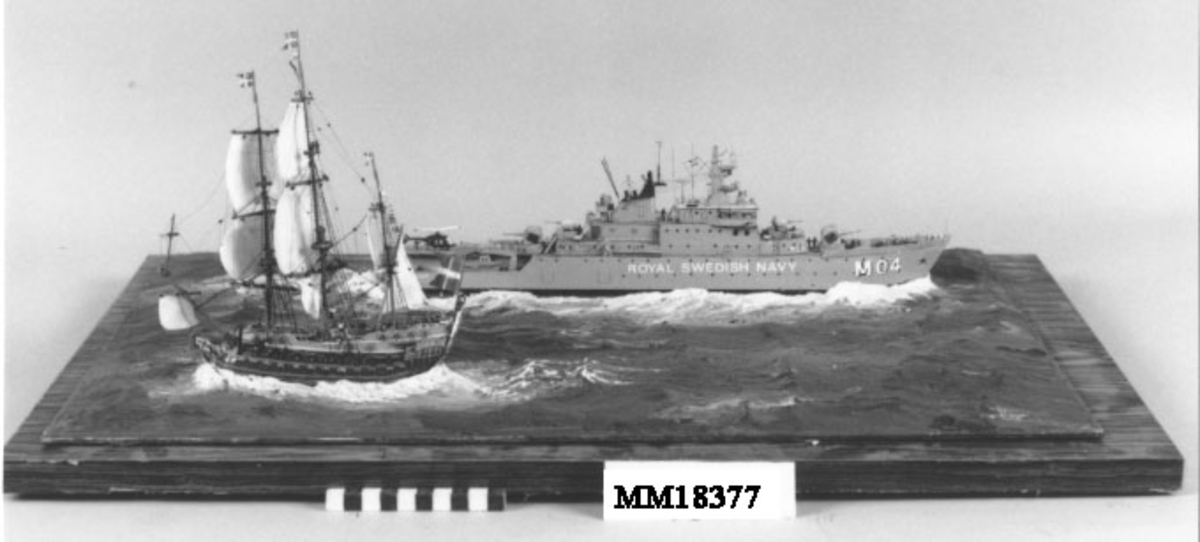 Fartygsdiorama bestående av två fartyg som passerar varandra: skeppet Karlskrona och minfartyget Carlskrona. Skeppet Karlskrona har tre master med satta segel, dubbla batteridäck, hög akter med fönster till överkajutan, akterspegeldekor bestående av stora riksvapnet, i fören krönt galjonsbild. Örlogsflaggor i masttopparna. Brunt skrov, överbyggnaden blå och gul. Minfartyget Carlskrona är grått, försett med helikopterplatta på akterdäck. Namn på båda sidor om fören "M 04 CARLSKRONA" samt "ROYAL SWEDISH NAVY" (Denna benämning realiserades aldrig ). På fartyget finns små figurer på däck och i master. Fartygen är placerade på rektangulär platta i ett hav av plastelina i gröna toner, signerad i ena hörnet i rött: " H.Biärsjö ". Försedd med glashuv med text: " 1680 Karlskronavarvet  1980 300 år ".