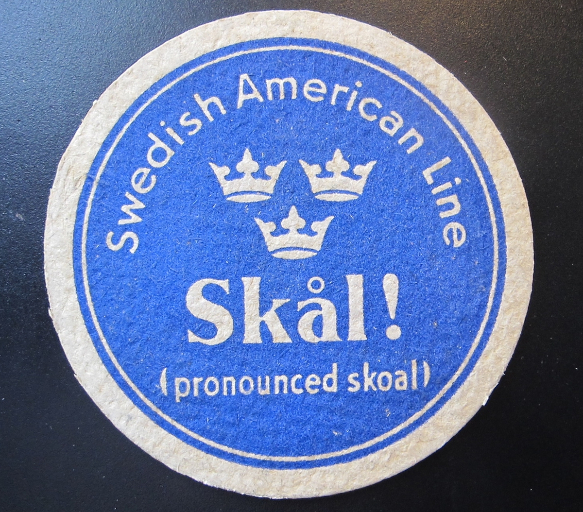 Runt glasunderlägg i beigevit papp med blått tryck, text i beigevitt: "Swedish American Line" "Skål!" "(pronounced skoal)" SAL:s emblem med tre kronor i mitten.