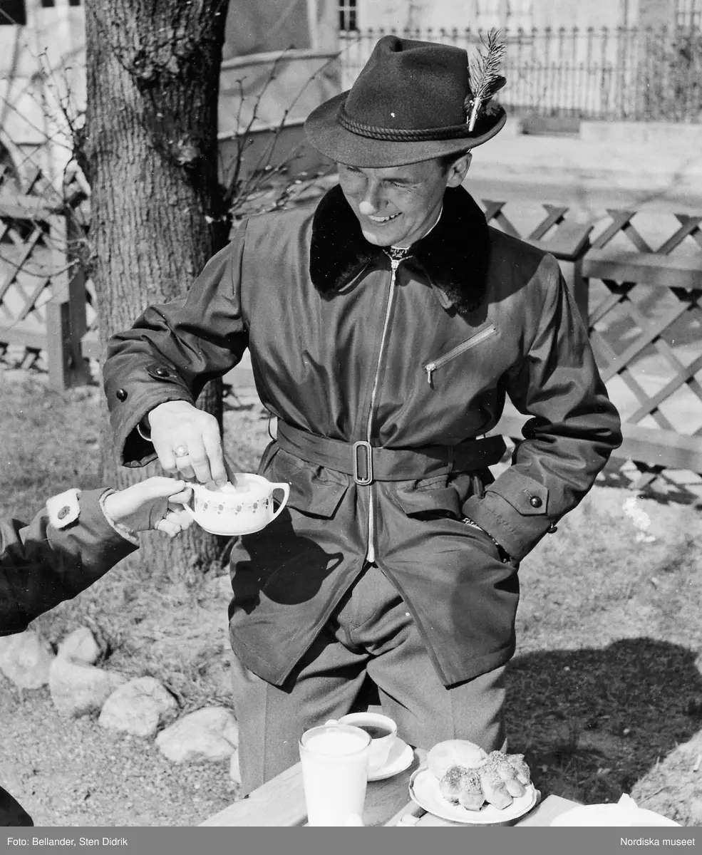 Modell i jacka med pälskrage och hårfiltshatt, tar socker ur en sockerskål.