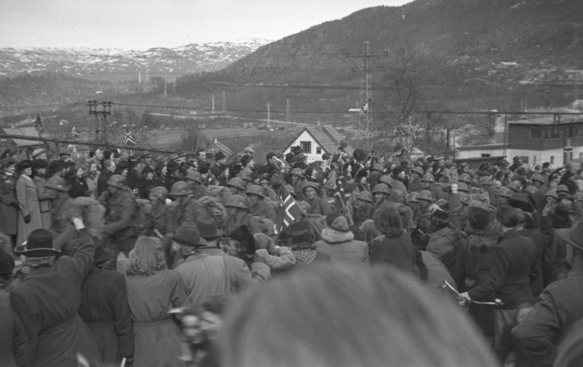 Folkemengde oppsamlet ved Narvik Jernbanestasjon. Soldater (polititropper) masjerer fra jernbanestasjonen, 8. mai 1945