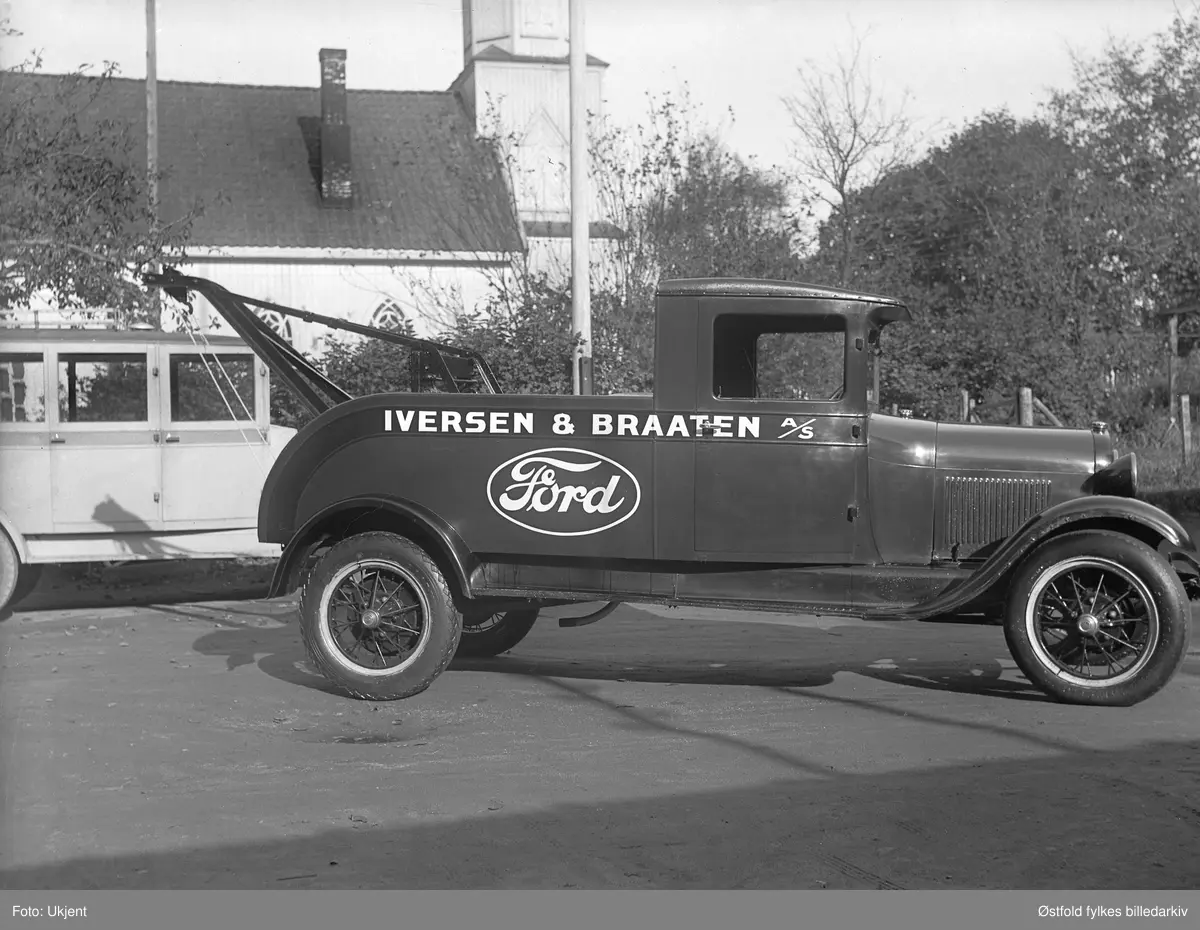 Service-vogn eller kranbil, tilhørende firmaet Iversen og Braaten A/S, fotografert på Mysen i Eidsberg ca. 1928-30. Bilden er en Ford A 1928-29 modell kranbil. 
En liten buss som skimtes i bakgrunnen. Bemerk kranbilens blankslitte høyre fordekk.