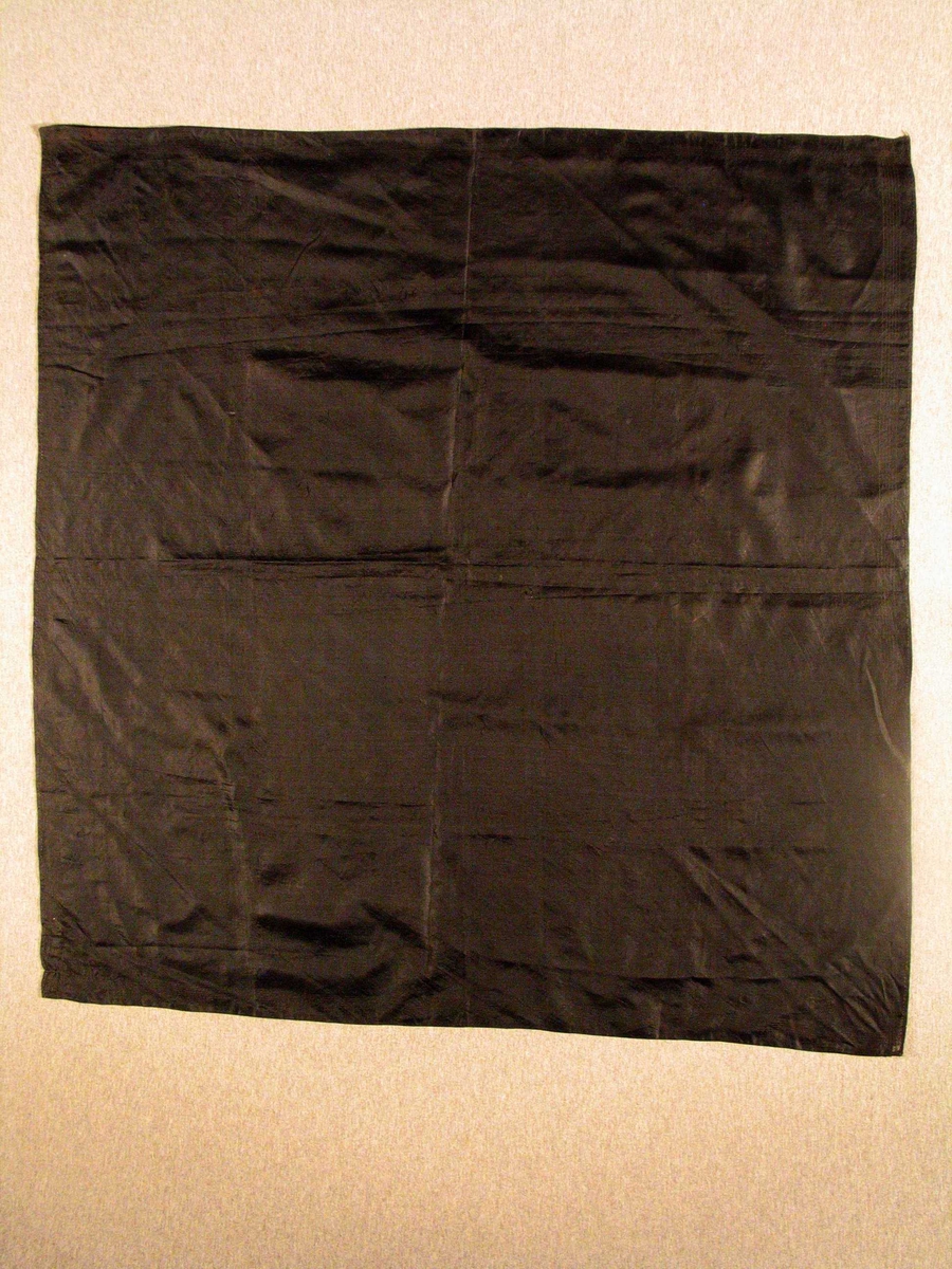 Rektangulær klut i svart silke. Kluten er håndsydd og det er en bord i svart. 