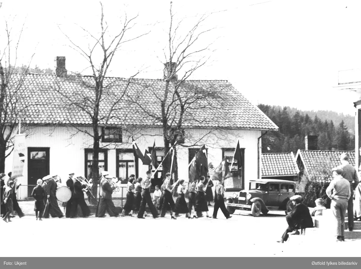 1.mai feiring i Ørje i Marker på 1940-50-tallet.
Foran i toget går ungdommer som bærer flagg med sigd, deretter følger et korps, før den første parolen. 
På trappen i forgrunnen av bildet sitter og står det skuelystne.

Bak toget synes Høiegården Amerikansk bil 1926-28 modell, amerikansk.