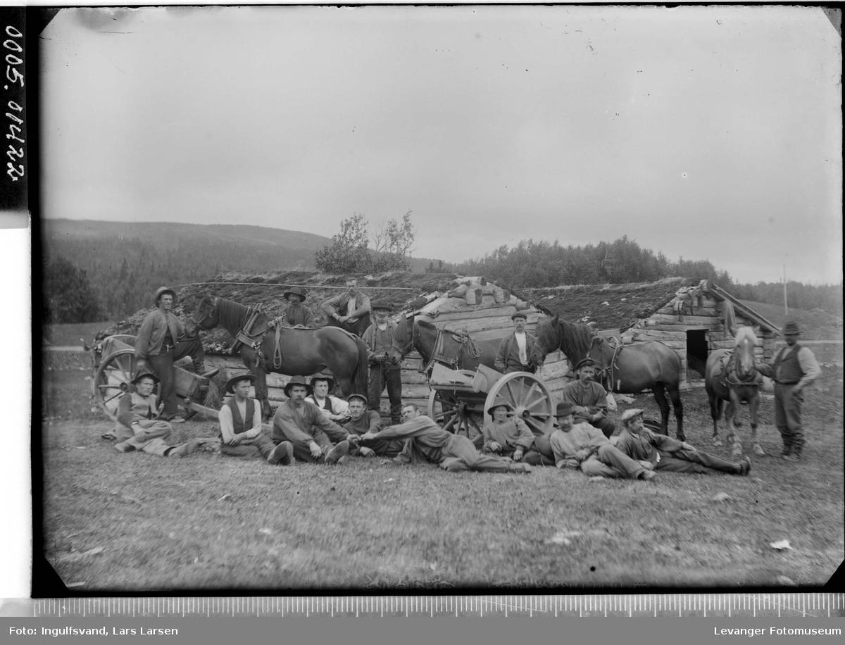 Gruppebilde av menn, hester og bikkkjærrer foran løer med torvtak.