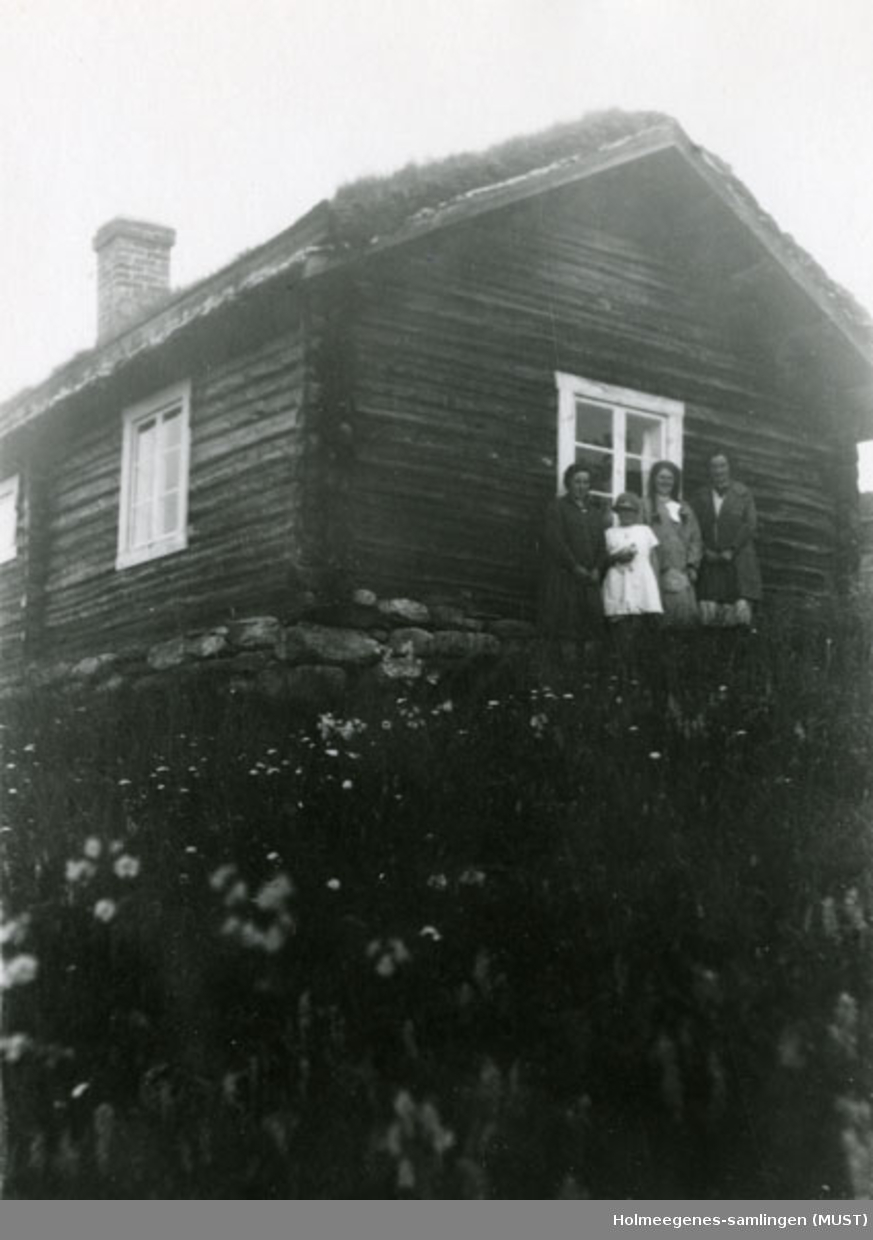 To kvinner og to jenter står foran en liten tømmerstue med torvtak og murpipe. Se også ST.K.HE 2007-011-0111 til -0126.