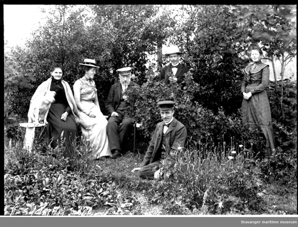 Arrangert foto av mennesker i hage (se ST.S 1981-011-0207), to damer og en mann sitter på en benk, en mann med panamahatt står bak en ripsbusk og foran sitter en mann med seilerlue. Til høyre i motivet står Julie Lund.